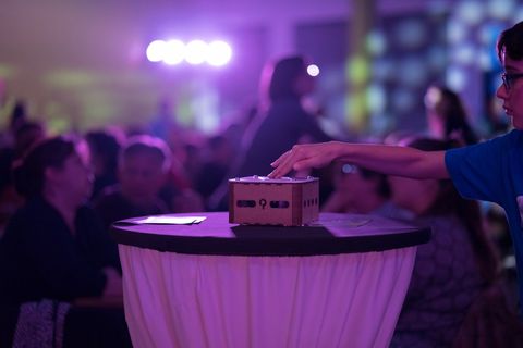 Une main appuie sur un buzzer Quizz Factory posé sur une table haute, la salle en fête à l'arrière-plan