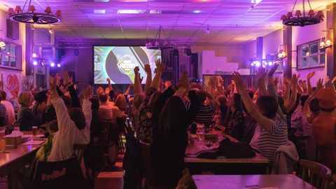 Une salle comble, bras levés, face à deux écrans de quiz sous des lumières violettes